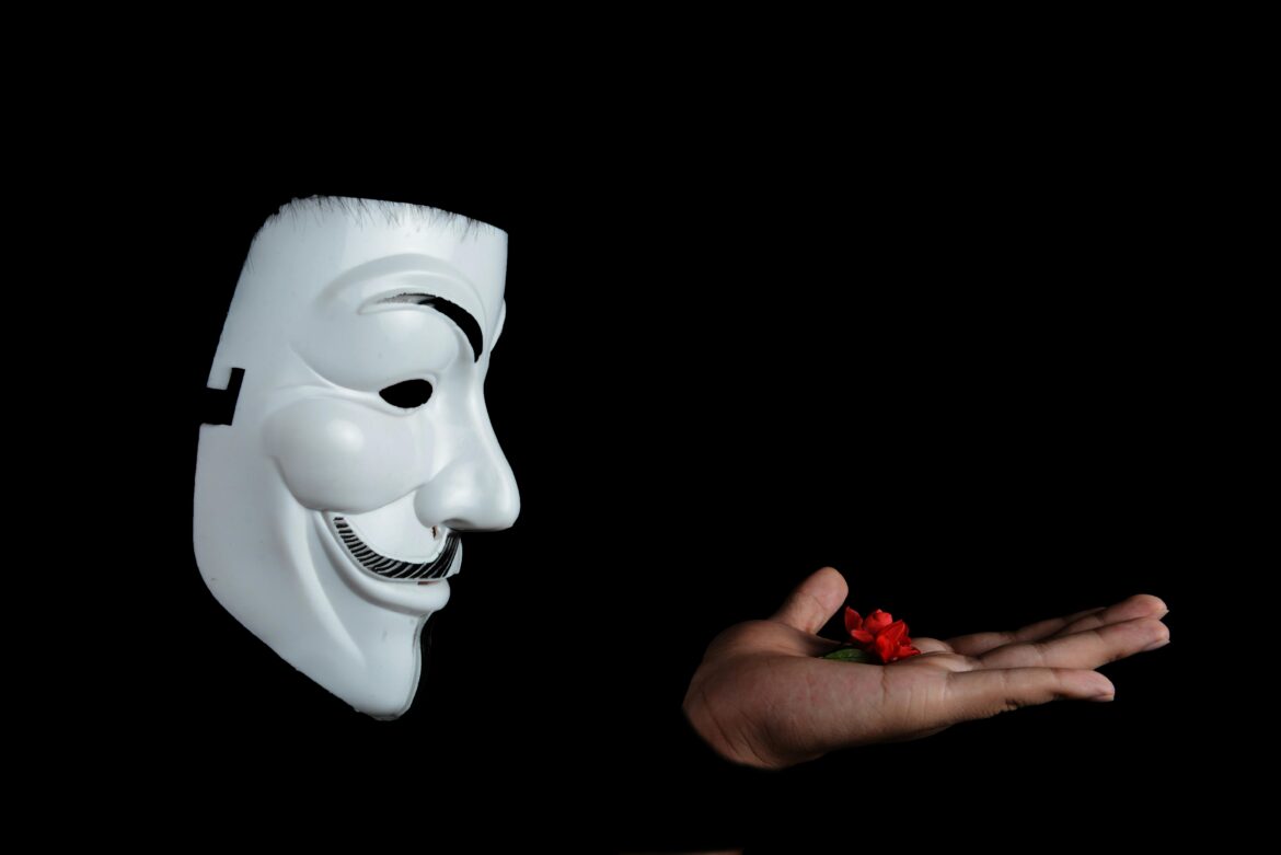 Side view of an anonymous mask facing a hand holding a red flower against a black background.